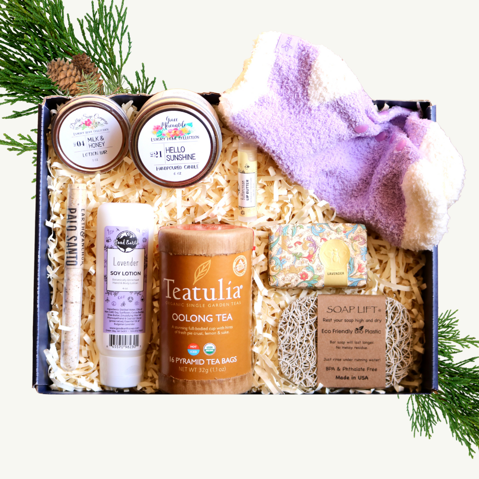 A gift basket containing various spa products including Teatulia organic tea, essential treatment socks, lip butter, hand and body lotion, a lotion bar, soap, and a candle, all arranged in a navy blue box with decorative greenery.