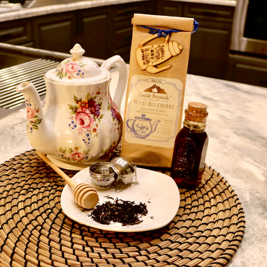 Cascade Botanicals Wild Blueberry Black Tea package displayed with a floral teapot, honey, and loose leaf tea accessories
