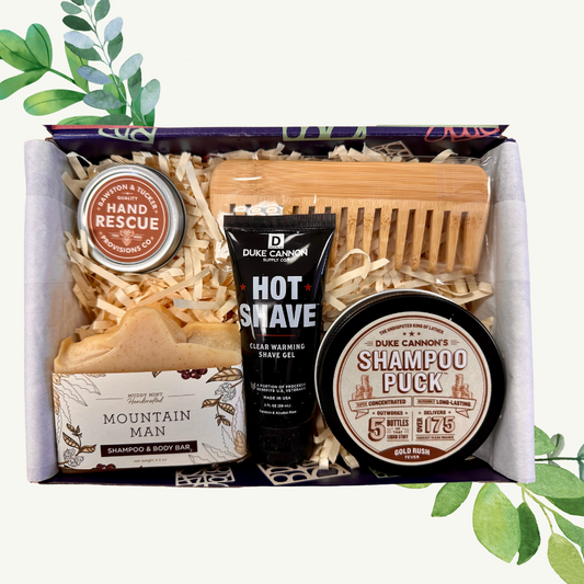 A grooming kit for men containing shampoo, body bar, shaving cream, and a wooden comb, packaged in a box with a mountainous design.