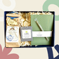 A gift set box containing a journal, a gold ballpoint pen, tea infuser, loose tea, and eye gels, presented against a white background with decorative flowers.
