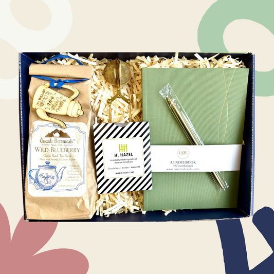 A gift set box containing a journal, a gold ballpoint pen, tea infuser, loose tea, and eye gels, presented against a white background with decorative flowers.