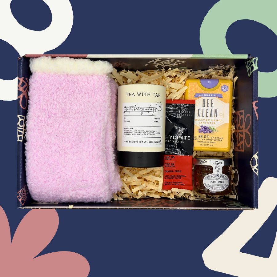 Gift box with various items including a pink fuzzy blanket, tea, and snacks on a colorful abstract background.