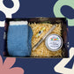 Box with blue fuzzy socks, shredded paper, and a candle labeled 'Hello Sunshine' on a colorful abstract background.