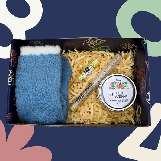 Box with blue fuzzy socks, shredded paper, and a candle labeled 'Hello Sunshine' on a colorful abstract background.