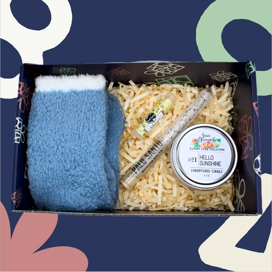 Box containing a blue fuzzy item, shredded paper, and a candle labeled 'Hello Sunshine' on a colorful background.