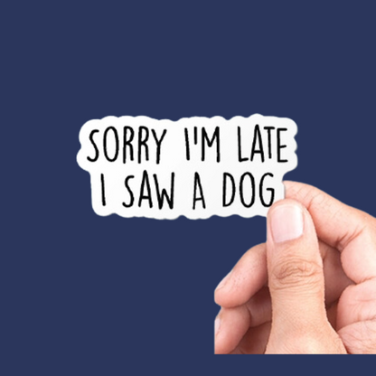 A sticker with the text 'Sorry I'm late I saw a dog' and a small illustration of a dog. The sticker is being held by a hand against a navy background.