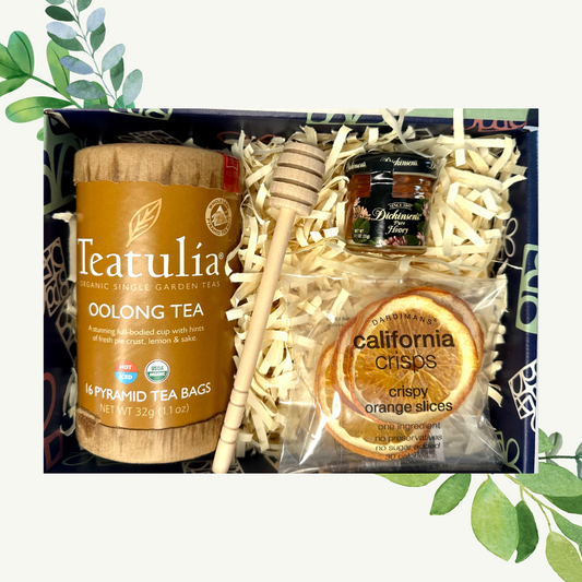 A gift basket containing Teatulia Organic Oolong Tea, Dickinson's Pure Honey, a honey dipper, and crispy orange slices, all packaged in a box with green leaves and a bee on the honey label.
