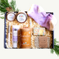 A gift basket containing various spa products including Teatulia organic tea, essential treatment socks, lip butter, hand and body lotion, a lotion bar, soap, and a candle, all arranged in a navy blue box with decorative greenery.