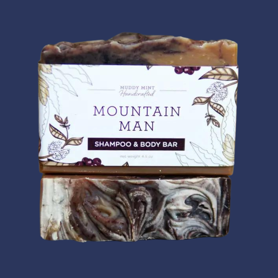 A bar of shampoo and body bar with a label that reads 'MUDdy Mint HANDCRAFTED', 'Mountain Man', 'Shampoo & Body Bar', and indicates the scent as Cedarwood Atlas, Fir Needle, Juniper Berry + Patchouli.
