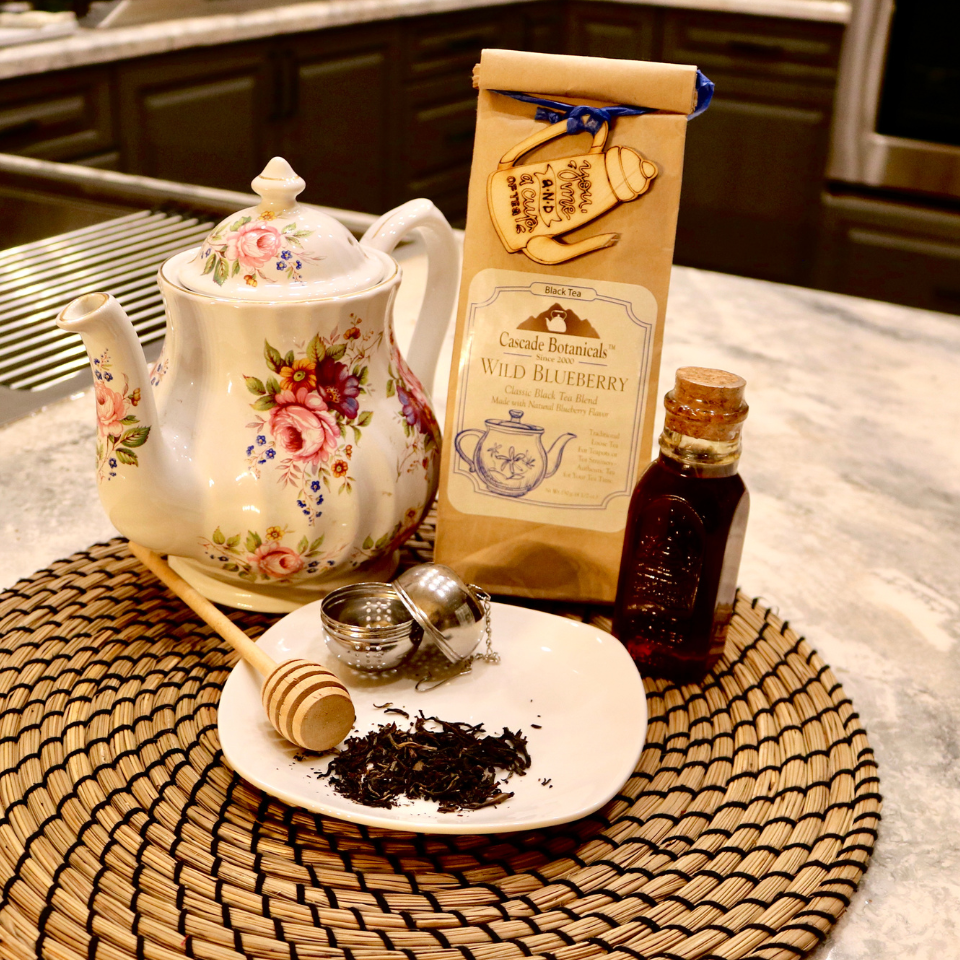 Cascade Botanicals Wild Blueberry Black Tea package displayed with a floral teapot, honey, and loose leaf tea accessories